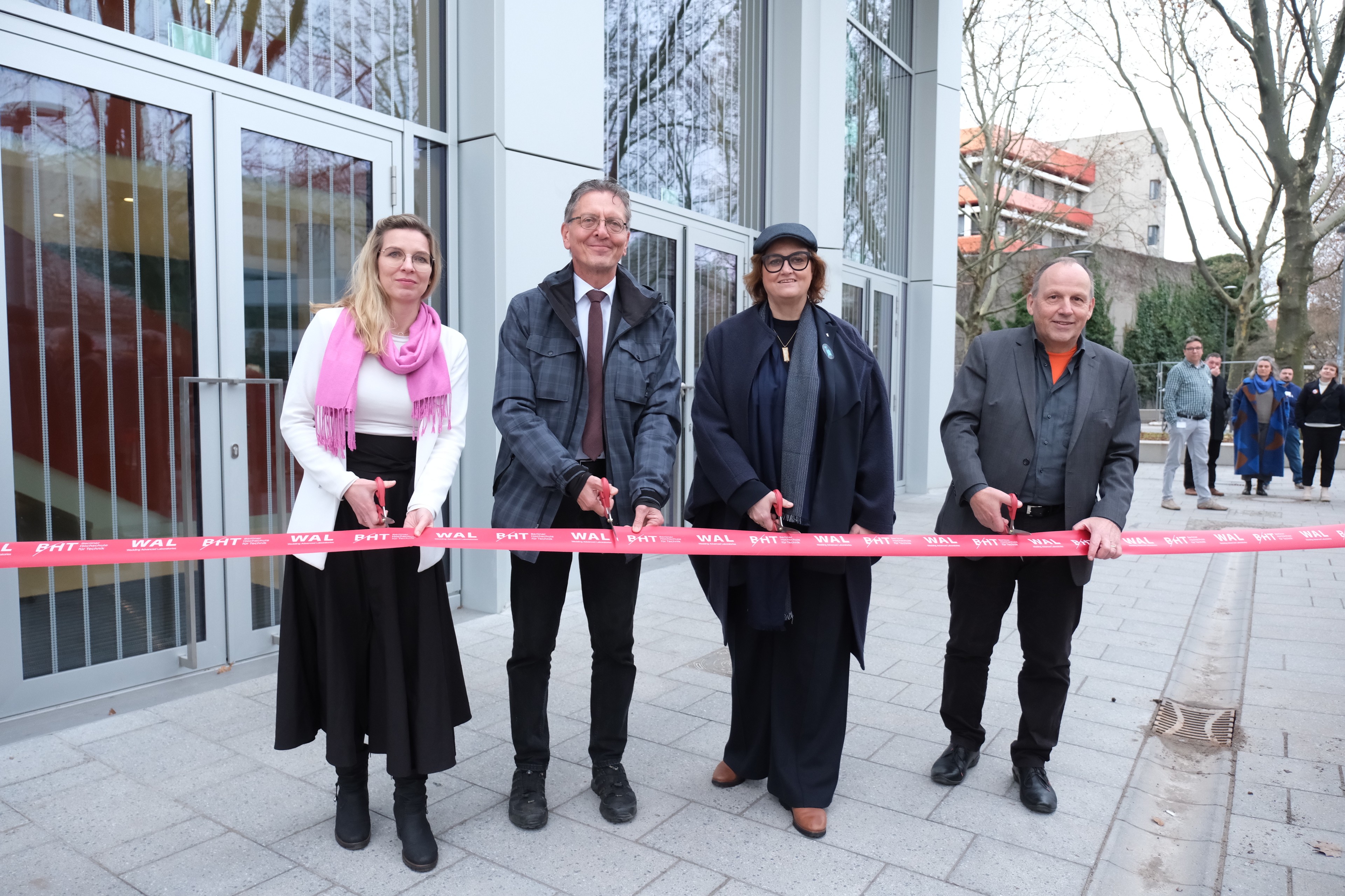 Vier Personen stehen vor einem Gebäude und halten Scheren bereit, um ein rotes Band durchzuschneiden