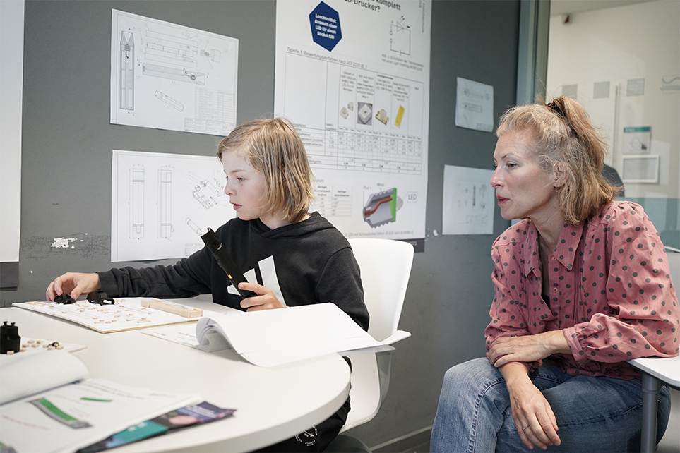 Ein Kind baut eine Taschenlampe aus dem 3D Drucker zusammen, die Mutter schaut dabei zu.