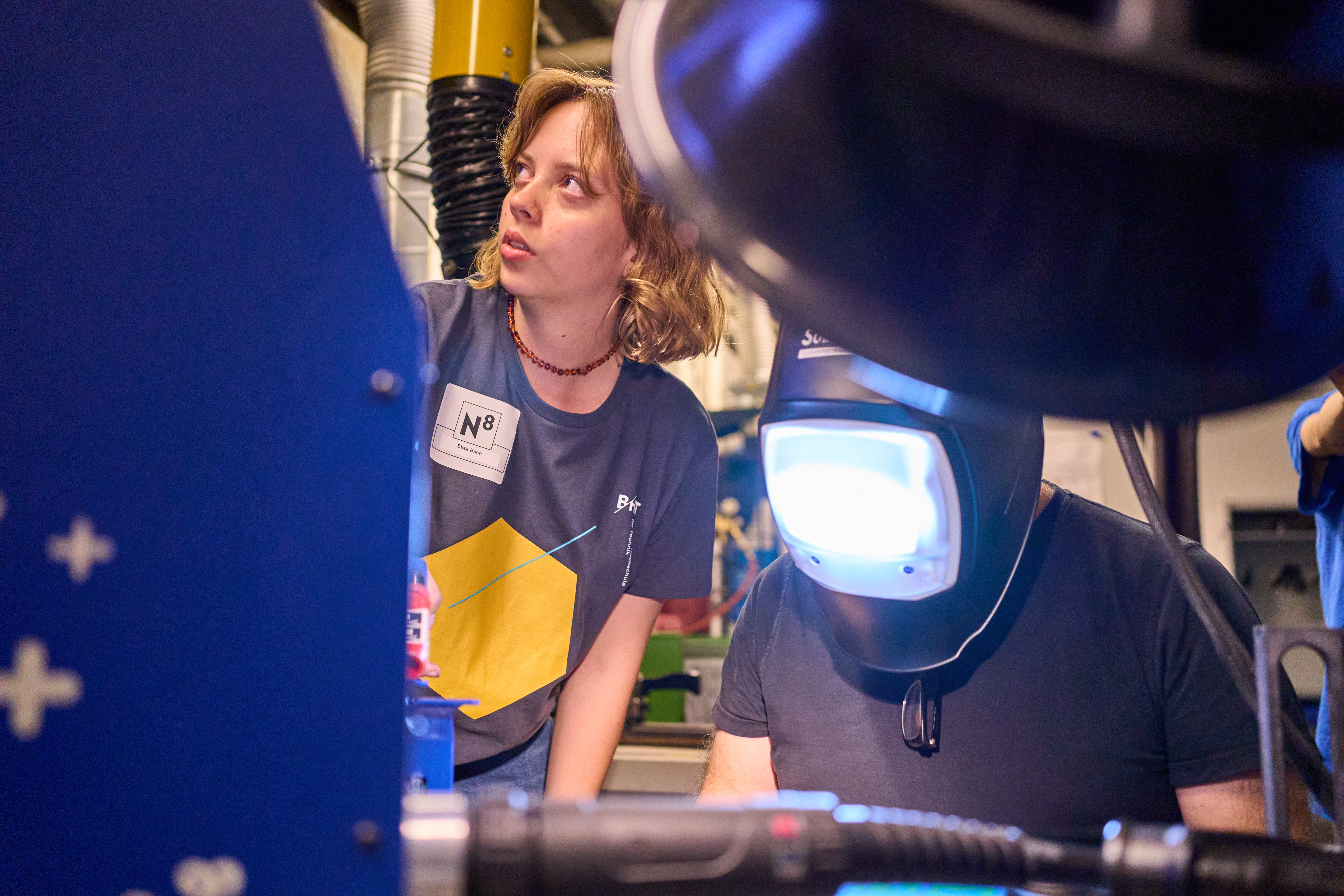 Eine Person trägt eine Schweißermaske, daneben steht eine Frau und erklärt etwas