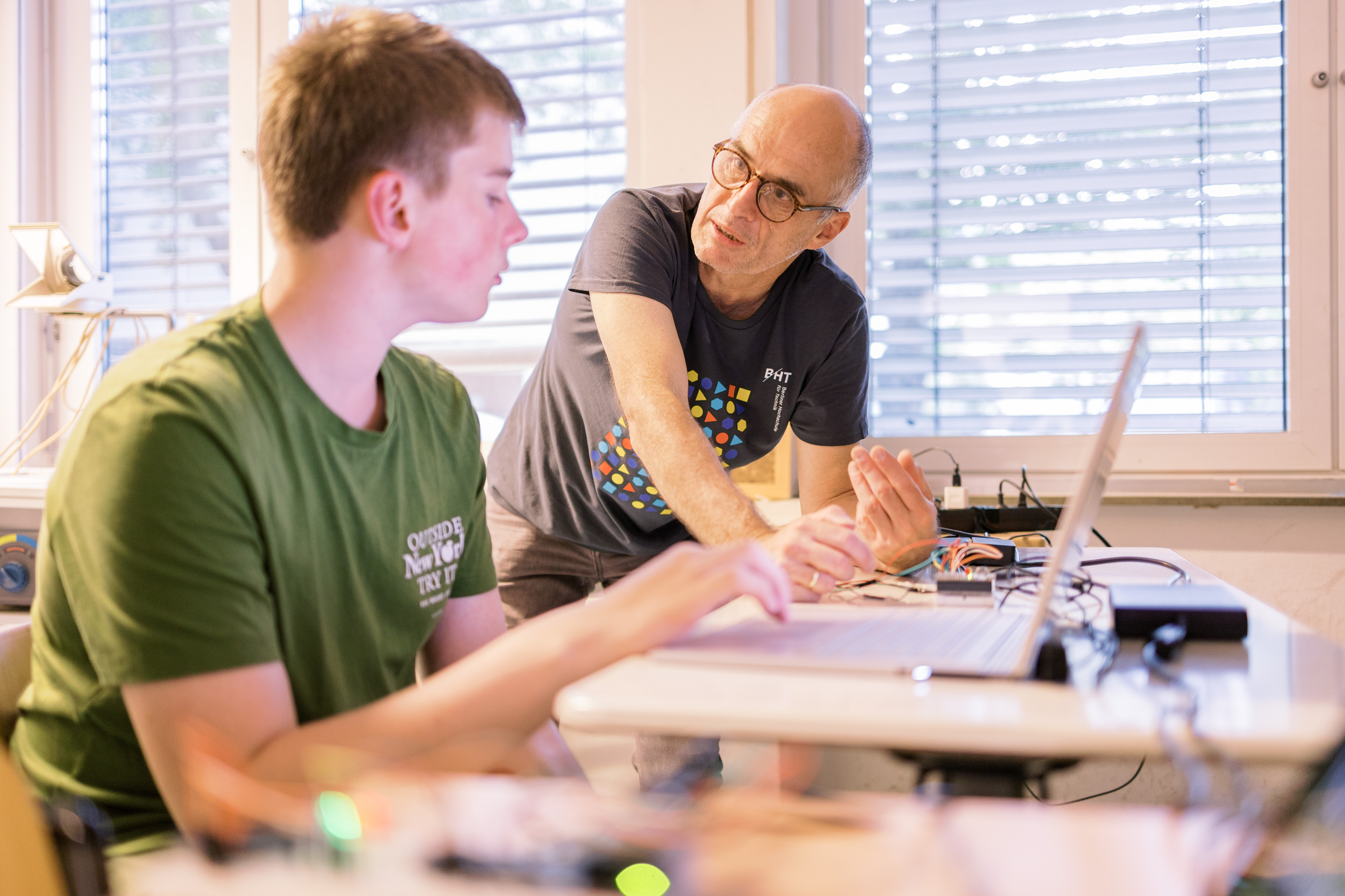 Ein Mann erklärt einem Jüngeren, der vor Laptop sitzt, etwas