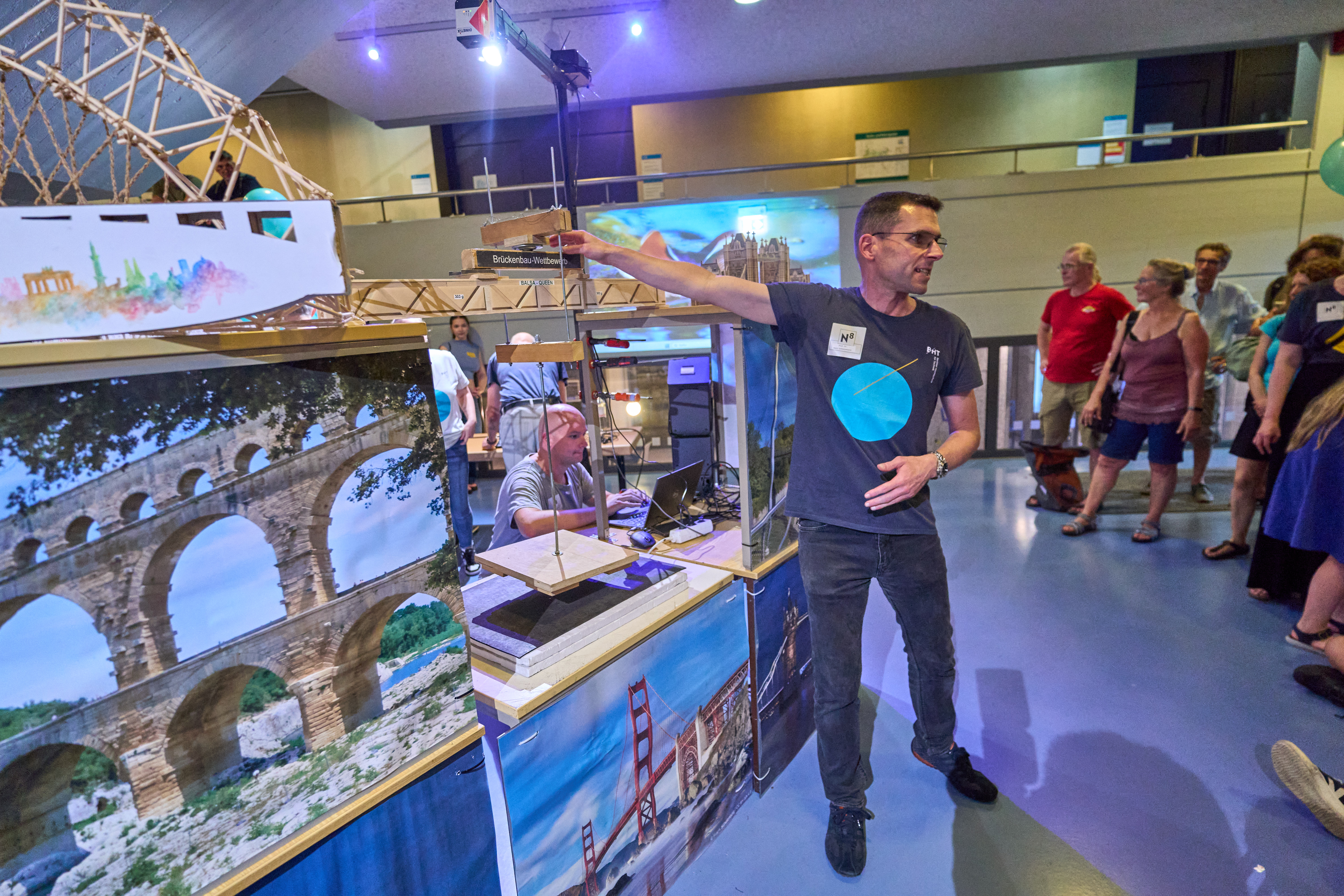 Mann steht an Brückenmodell und spricht zum Publikum