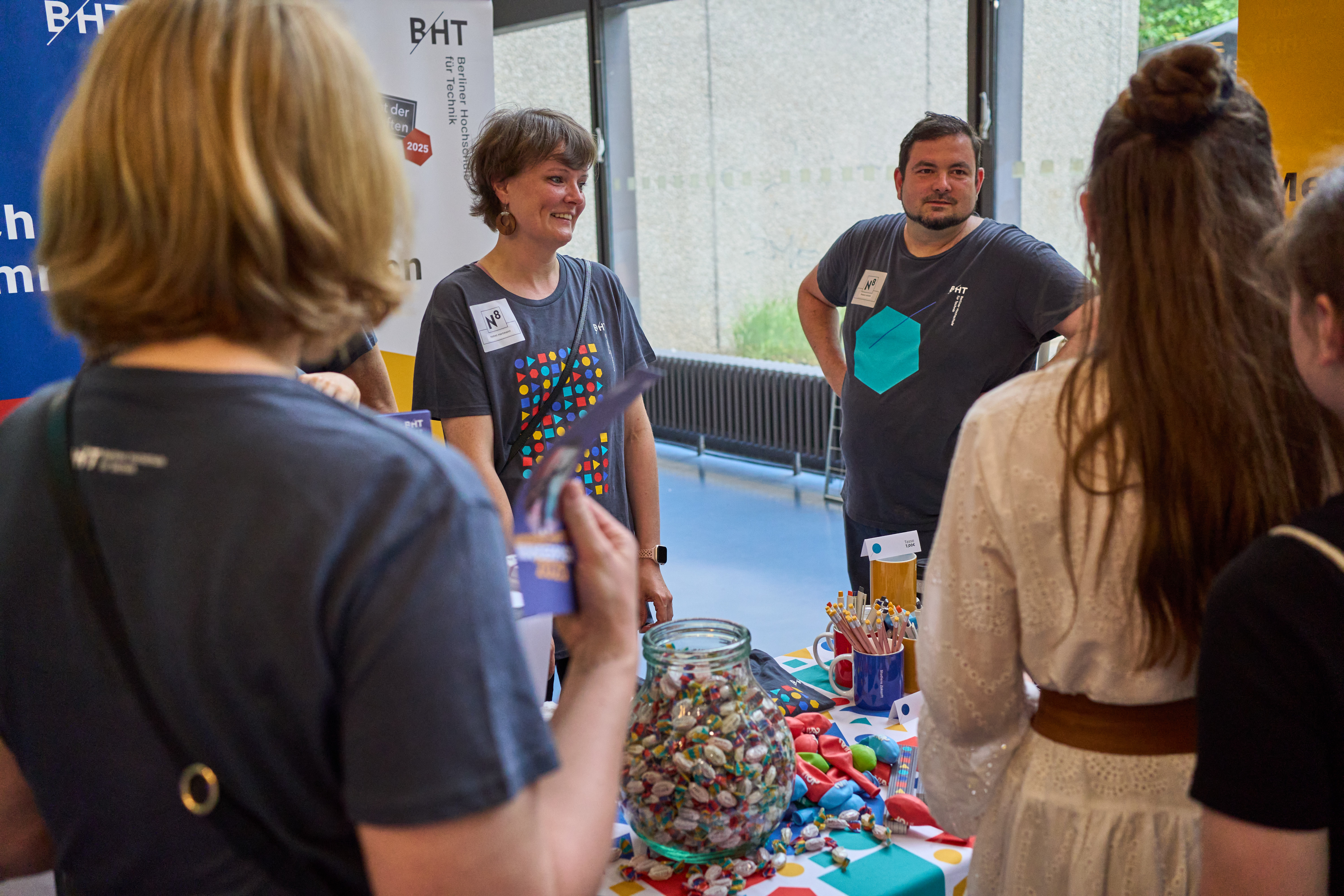 Mitarbeiter*innen begrüßen Besucher*innen an einem Stand.