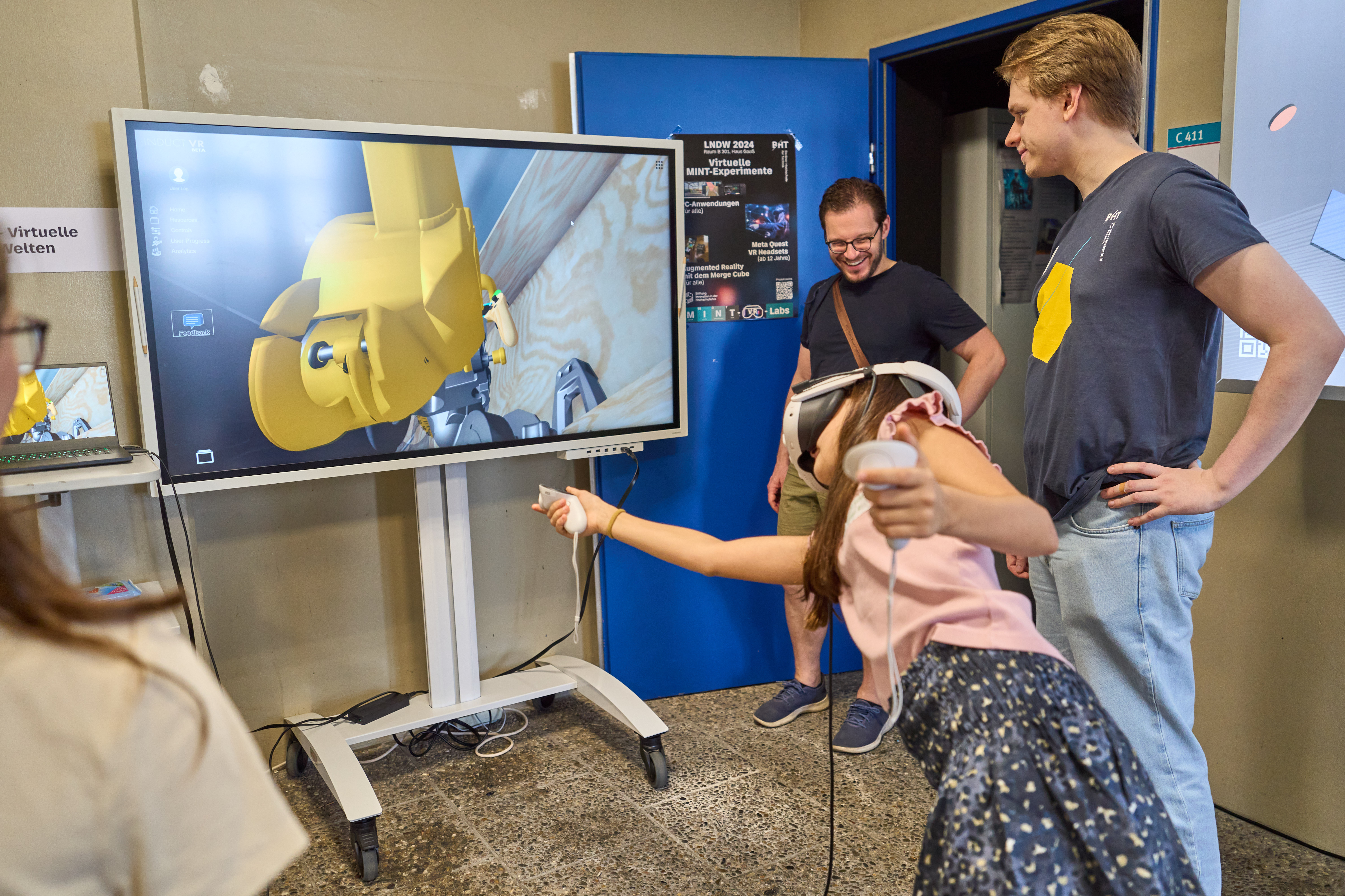 Mädchen mit VR-Brille interagiert, im Hintergrund Bildschirm und mehrere Personen.