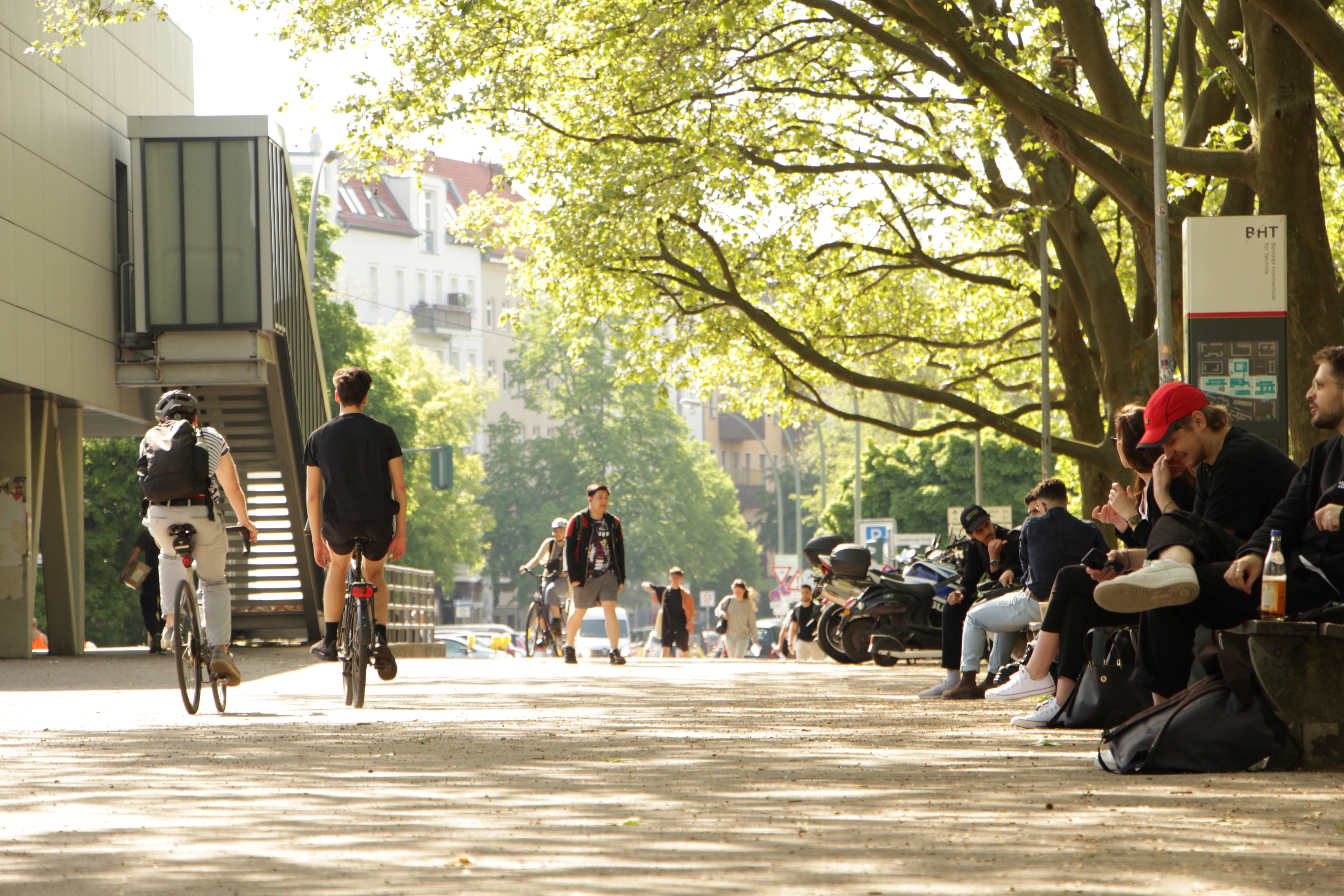 Menschen auf dem Campus der BHT, unter anderem zu Fuß und mit dem Rad unterwegs, im Hintergrund Autos und Motorräder 