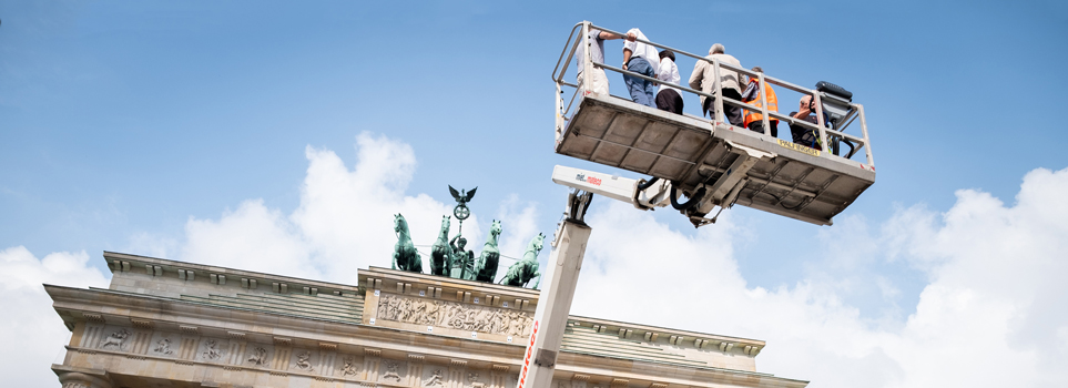 Relief des Brandenburger Tors vor blauem Himmel und davor in Menschen auf Hebebühne in großer Höhe